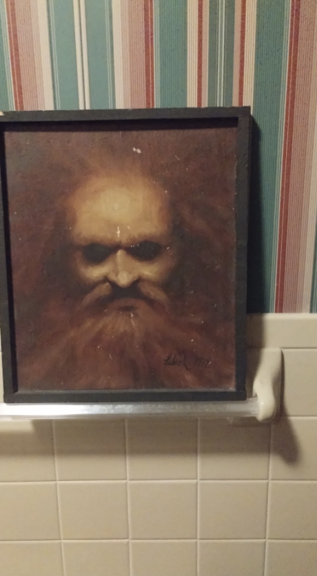 A framed painting of a bearded man's face with a solemn expression is mounted on a small ledge in a room with striped wallpaper and white tiled walls. The painting has a dark, moody tone.