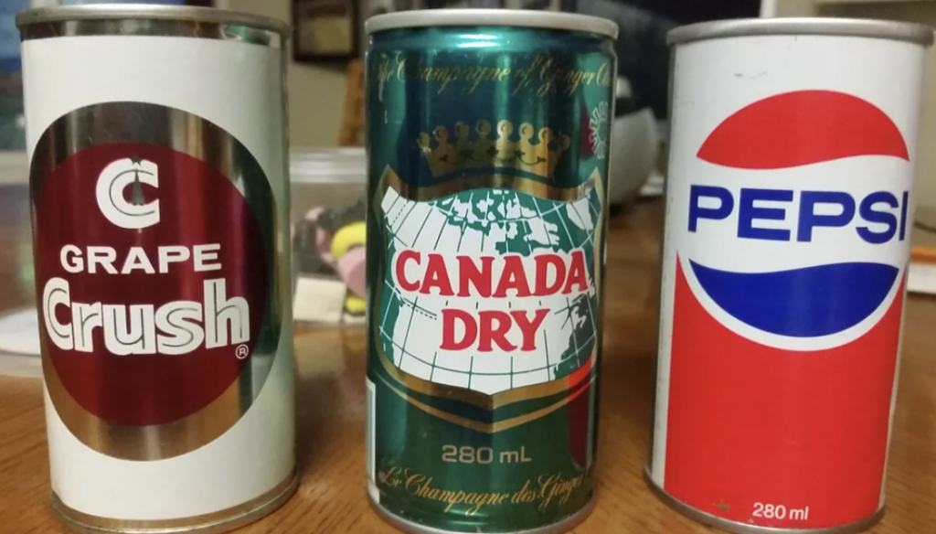 Three vintage soda cans are displayed on a wooden surface. From left to right: a Grape Crush can, a Canada Dry can, and a Pepsi can. Each can has distinct branding and design from an earlier era.