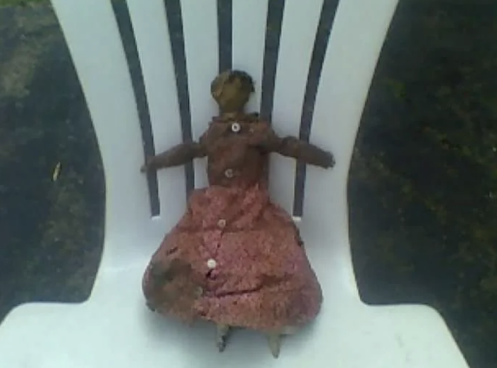 A worn-out, vintage doll dressed in a tattered red dress is propped up on a white plastic chair. The doll appears to have damage on its dress and legs, with some missing features and an overall weathered appearance. The background shows a dark, indistinct surface.