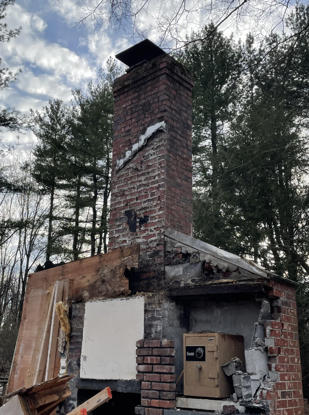 A brick chimney with visible damage is all that remains of a structure, surrounded by forest. The surrounding area is filled with debris and partially broken walls. The background features trees and a partly cloudy sky.