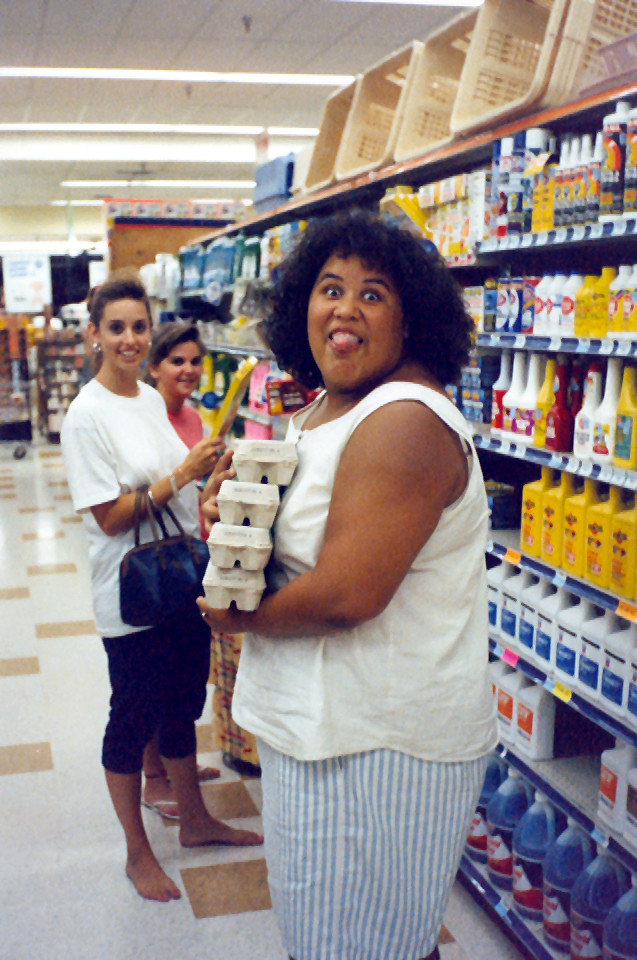 A woman in a white top and striped skirt makes a funny face while holding egg cartons in a grocery store aisle. Two women in the background laugh and look on, one holding a purse and the other a yellow item. The shelves are stocked with various products.