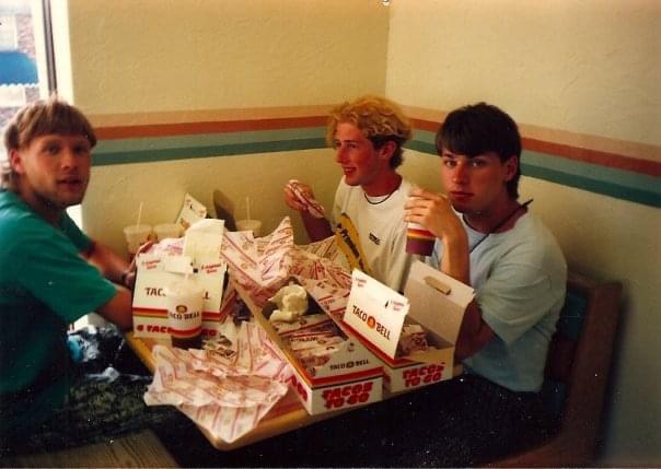 Three young men are seated in a Taco Bell restaurant booth with a table full of Taco Bell food packaging and drinks. They are casually dressed, and one of them is holding a drink cup. The restaurant features a simple, striped wall design.