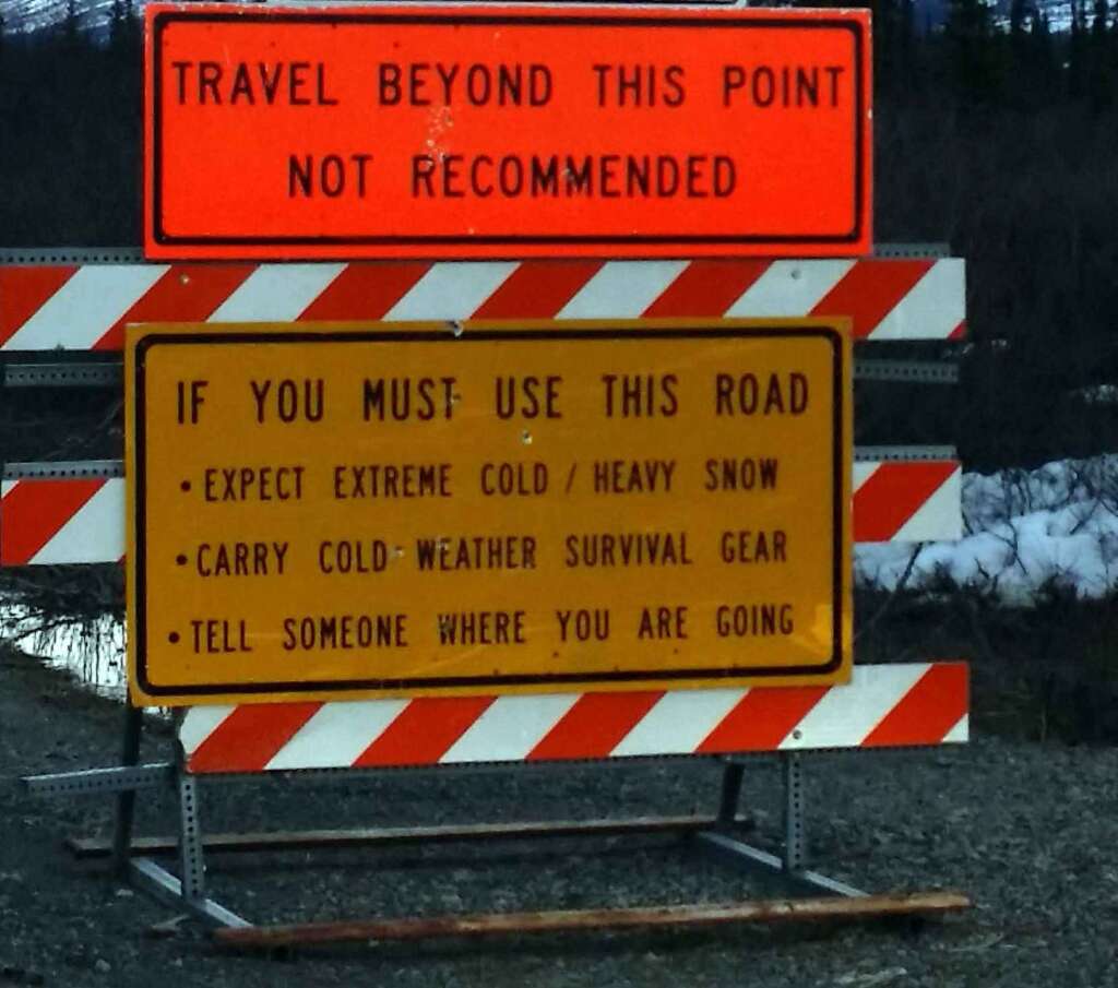 Two road signs are posted. The top sign in orange reads: "TRAVEL BEYOND THIS POINT NOT RECOMMENDED." The bottom sign in yellow reads: "IF YOU MUST USE THIS ROAD - EXPECT EXTREME COLD / HEAVY SNOW - CARRY COLD WEATHER SURVIVAL GEAR - TELL SOMEONE WHERE YOU ARE GOING.