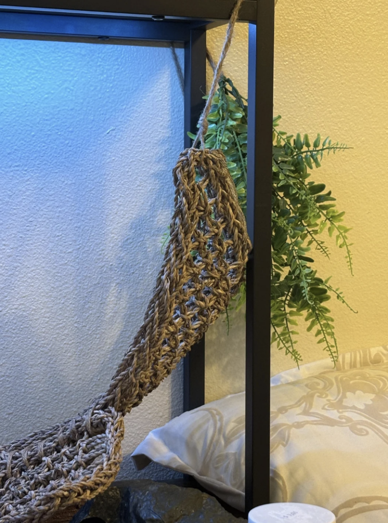 A small hand-knitted hammock hangs from a shelf with a leafy green plant in the background. Below the hammock, a pillow with beige floral patterns lies on a grey surface, and a white cylindrical container is placed nearby.