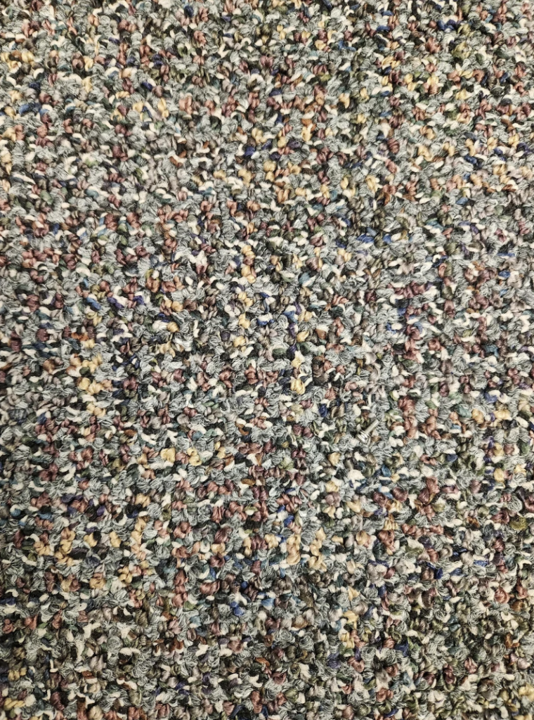 A close-up view of a textured carpet with a multi-colored pattern consisting primarily of shades of green, brown, blue, and hints of yellow and red. The dense, looped fibers create a rough, tactile surface.
