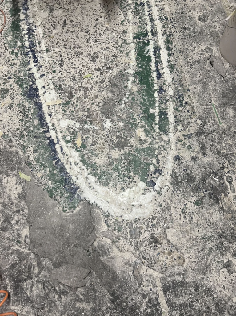 A textured gray surface, possibly concrete, with a faded green paint area outlined by white brushstrokes in a rough oval shape. There are scattered small debris and specks of white paint, giving the surface a worn and weathered appearance.