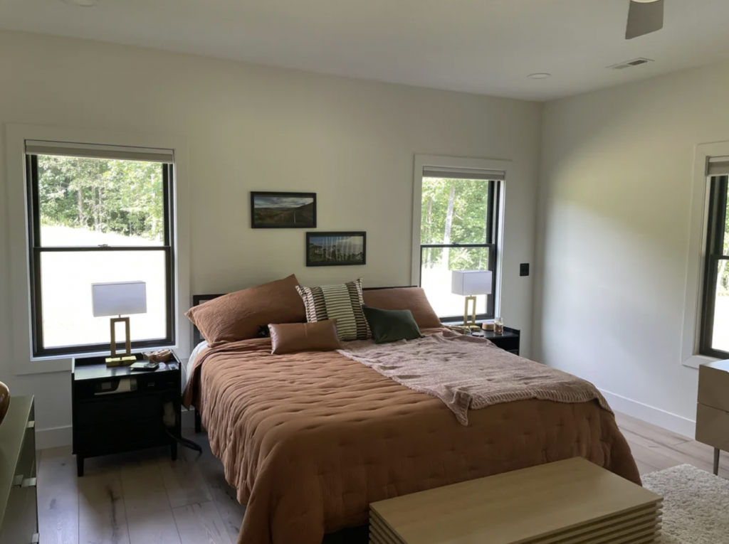 A cozy bedroom with large windows, a brown quilted bedspread, and decorative pillows on the king-sized bed. Two bedside tables with lamps and framed art above the headboard adorn the room. Natural light flows through the windows, highlighting the wooden floor.
