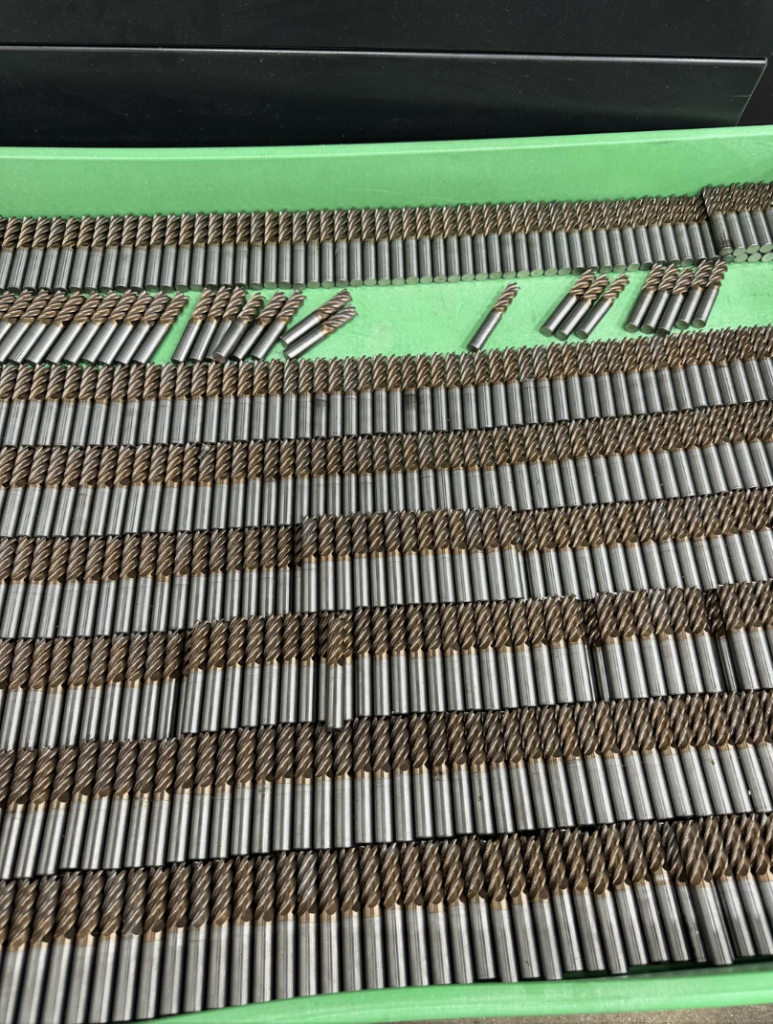 A collection of metal drill bits arranged neatly in rows on a green foam backing. The drill bits are of various sizes, with larger ones at the bottom and smaller ones at the top. The bits have cylindrical shanks and spiraled cutting edges.