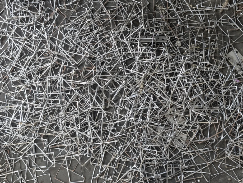 A large pile of twisted and tangled metal pieces, scattered across a concrete surface. The overlapping metal components create an intricate and chaotic pattern, with varying shapes and lengths intertwined amidst each other.