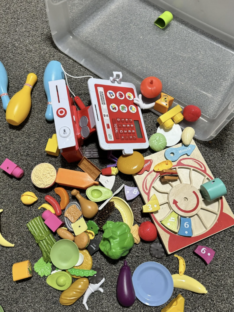 A collection of colorful plastic toy food items, a toy cash register, and bowling pins are scattered on a carpeted floor next to a clear plastic container. Items include fruits, vegetables, play money, and part of a board game.