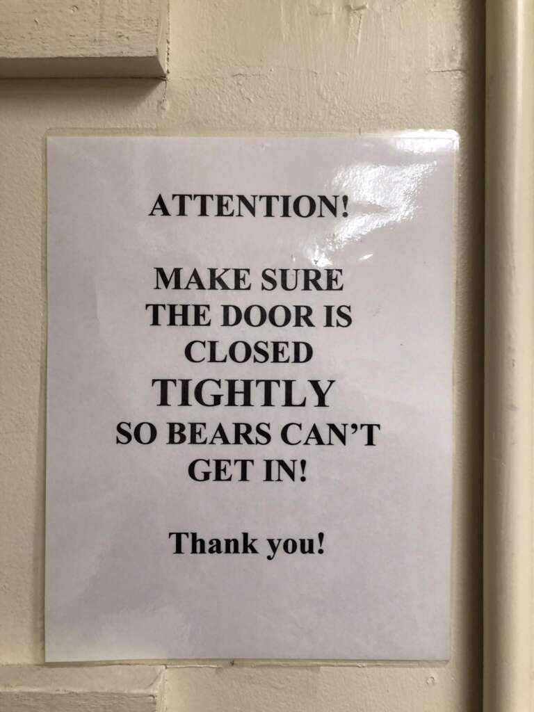 Sign on a door reads: “ATTENTION! MAKE SURE THE DOOR IS CLOSED TIGHTLY SO BEARS CAN'T GET IN! Thank you!” The sign is printed on white paper with black text and is taped to a pale-colored door.