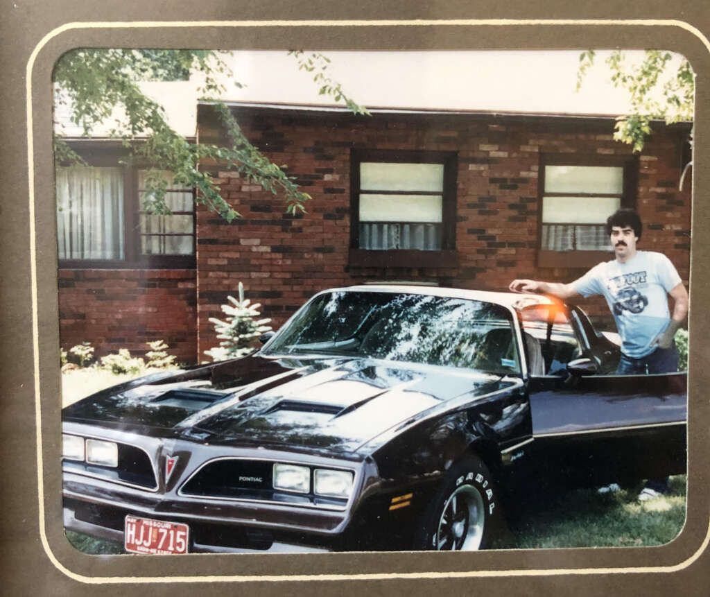 A man stands next to a black Pontiac Trans Am with one hand resting on the open driver's side door. The car is parked in front of a brick house with a few trees in the yard. The man is wearing a light blue T-shirt with an image on it.