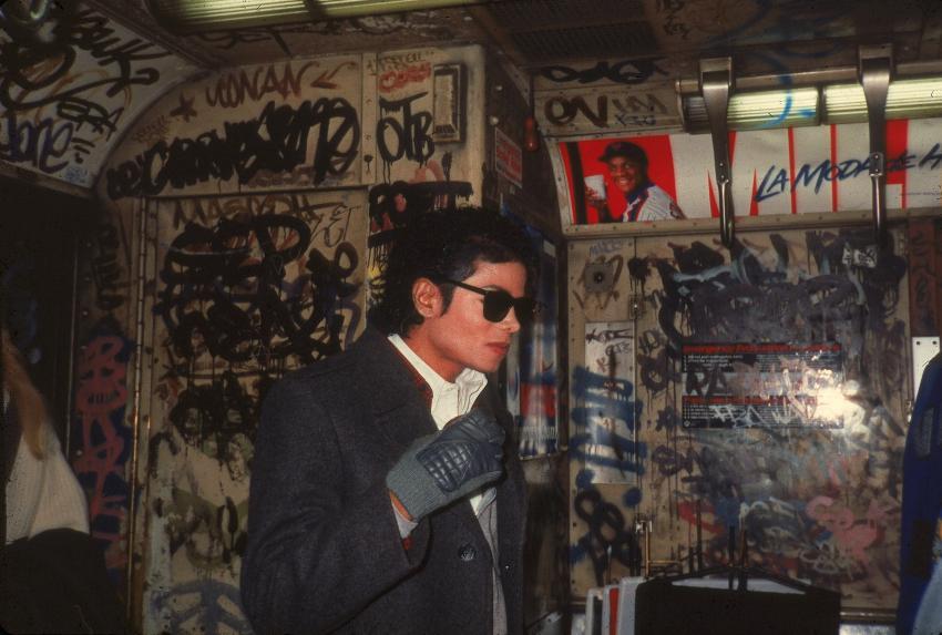 A person wearing sunglasses and a coat with gloves is standing in a dimly lit, graffiti-covered space. The walls are adorned with various tags and posters, including one with a smiling individual and vibrant colors. The atmosphere is urban and gritty.