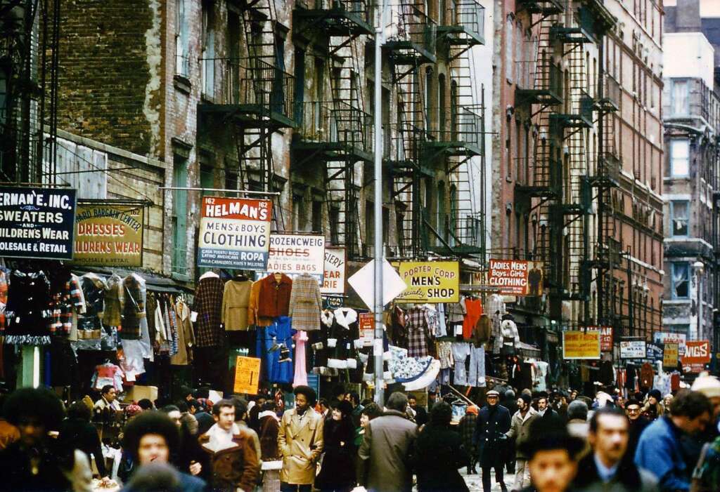 A busy city street scene with numerous people walking past storefronts. The buildings are adorned with signs advertising various shops, including clothing and hat stores. Clothes are displayed on racks outside the shops, and fire escapes are visible on the buildings.