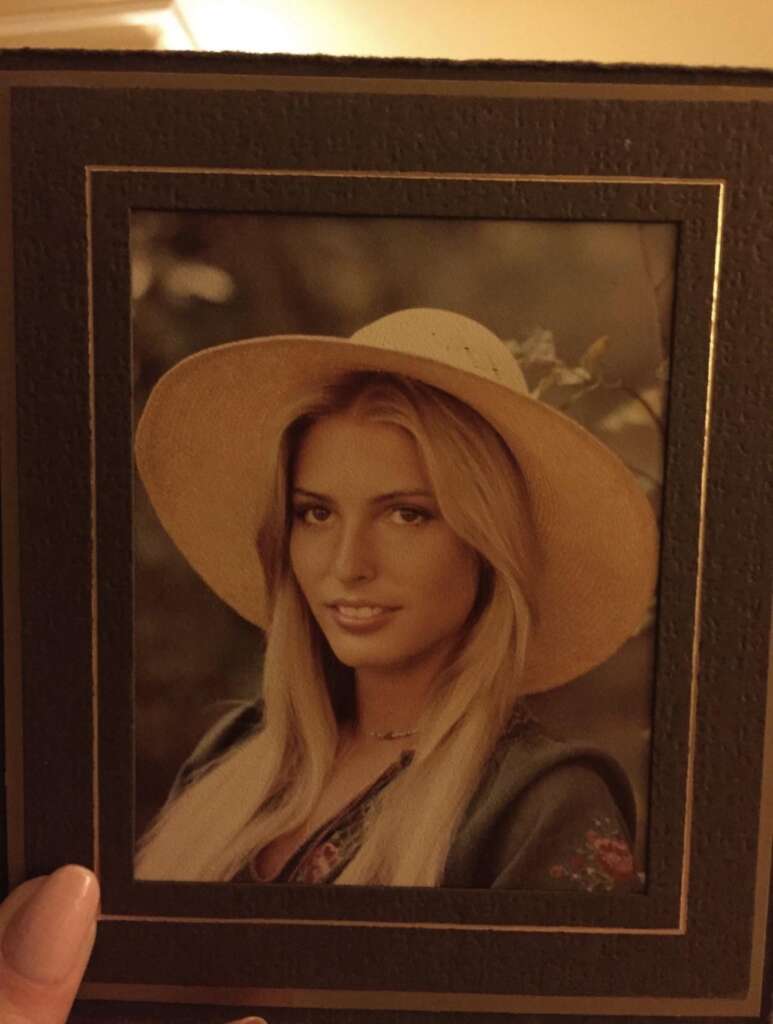 A framed portrait of a young woman with long blonde hair, wearing a wide-brimmed straw hat and a dark top with floral patterns. She is smiling subtly and looking directly at the camera. The background appears to be an outdoor setting with soft lighting.