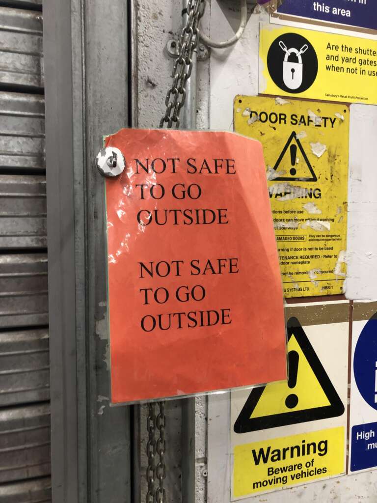 A bright orange sign reads "NOT SAFE TO GO OUTSIDE" in bold, all-caps lettering, hanging on a chain against a metallic wall. Various safety warning labels are visible in the background, emphasizing caution.