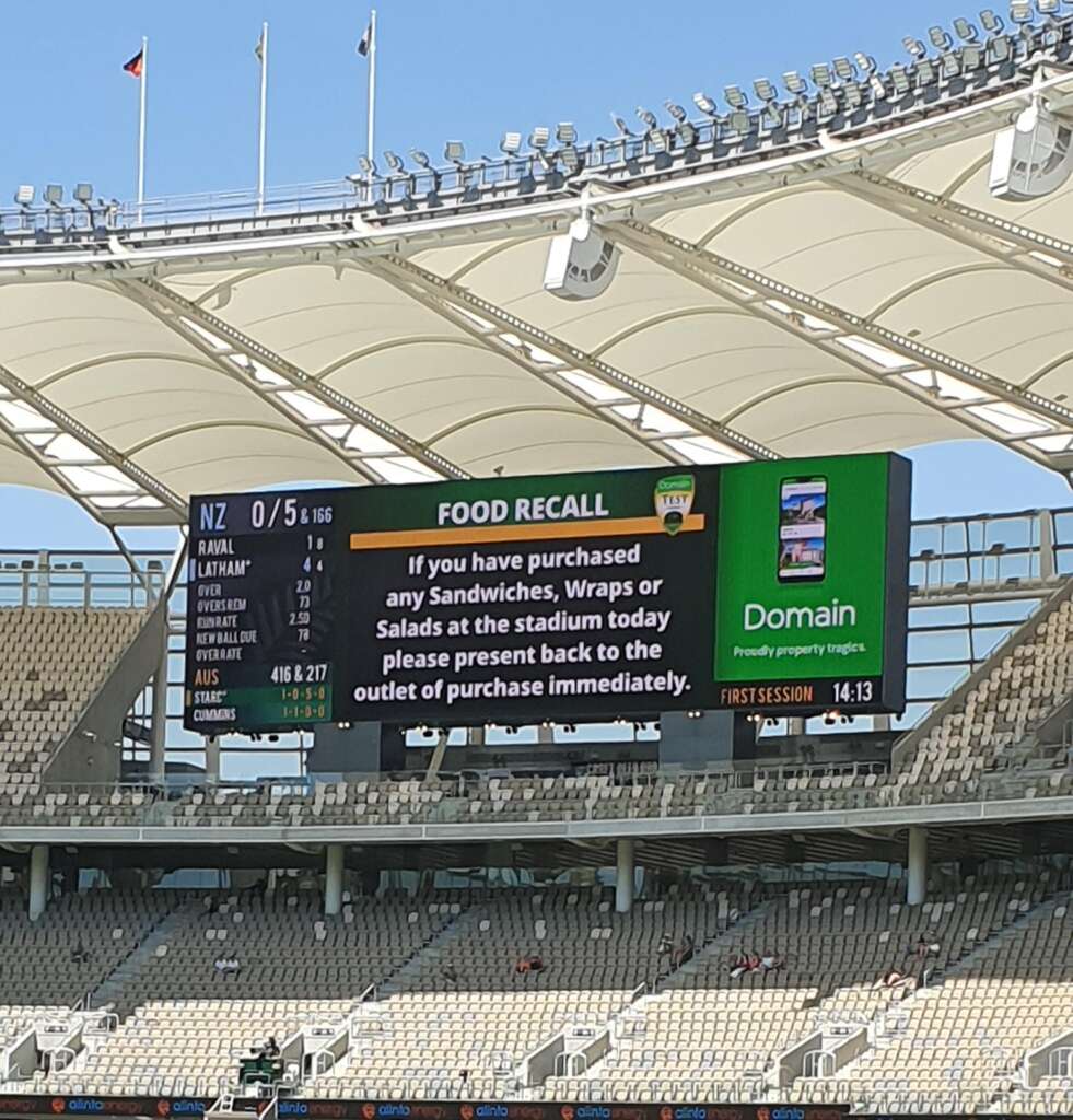 An empty stadium with a large screen displaying a food recall alert. The alert instructs not to consume sandwiches, wraps, or salads purchased at the stadium and to return them to the outlet. The scoreboard shows a cricket match between NZ and another team.