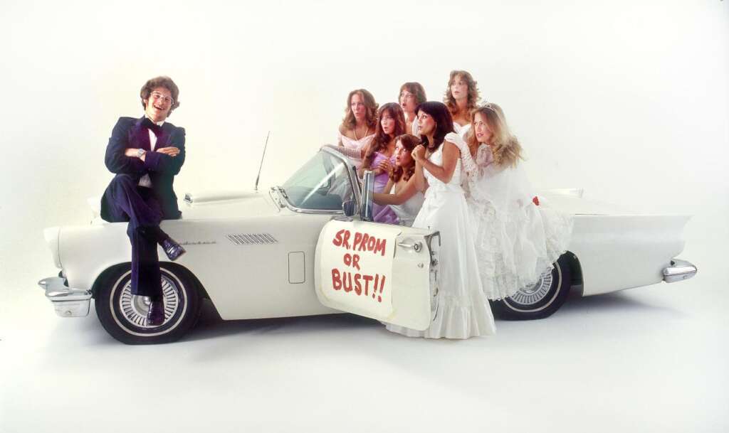 A group of seven young women in prom dresses pose around a white vintage convertible car. One young man in a suit sits on the driver's side door, which has a sign saying "SR. PROM OR BUST!!" The background is a plain white studio setting.