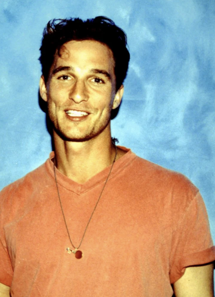 A man with short, wavy hair smiles at the camera against a light blue background. He is wearing a red t-shirt and a simple necklace with a pendant.