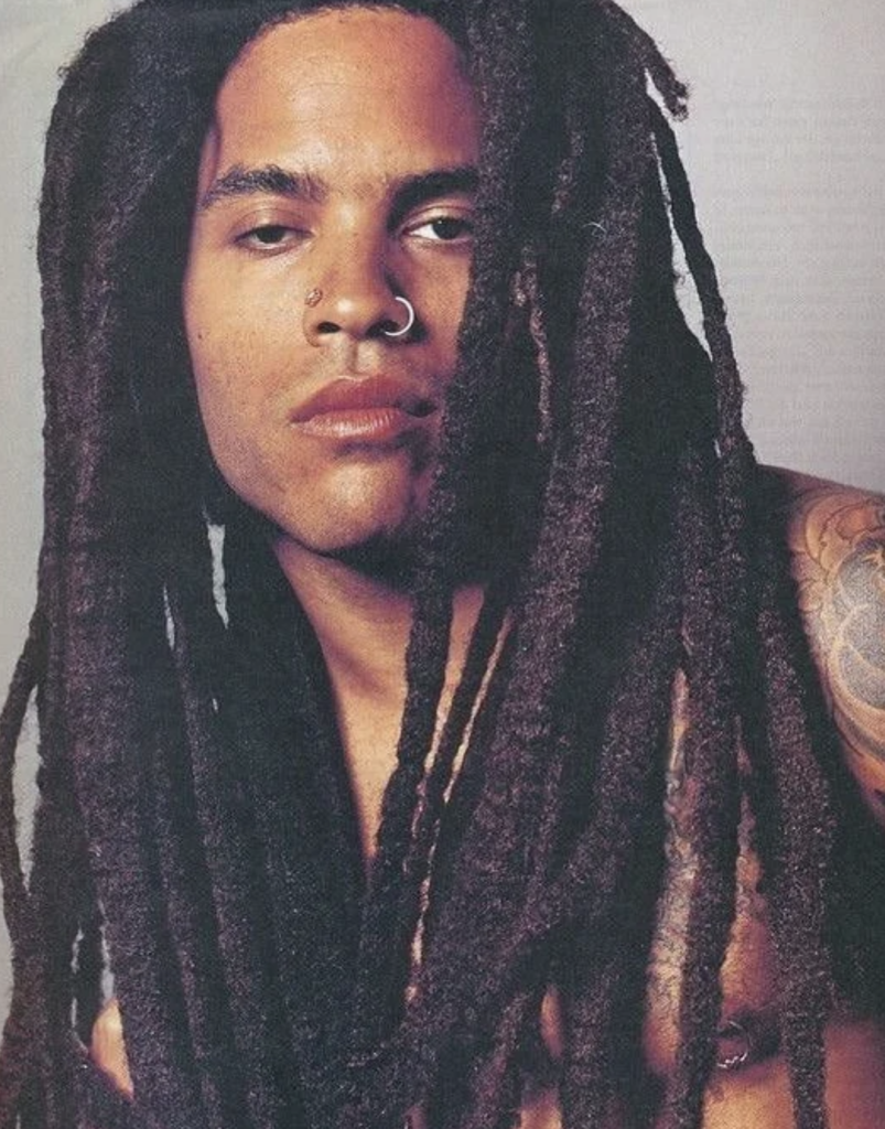A person with long, dark dreadlocks and a nose ring is portrayed in a close-up shot. They have a neutral expression and are shirtless, showing part of a tattoo on their upper arm. The background is plain and out of focus.