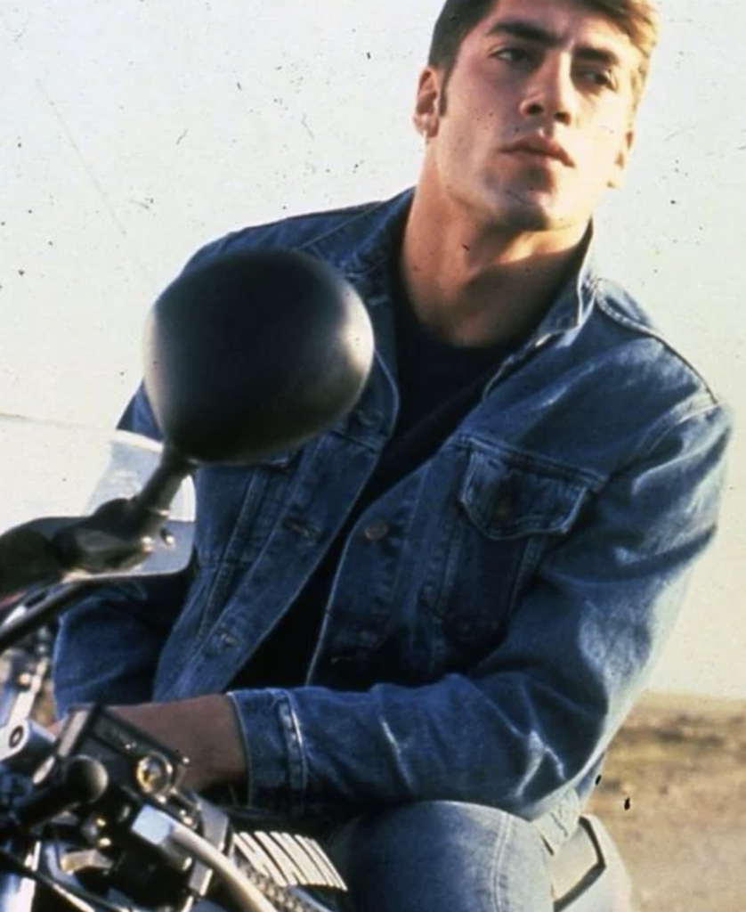 Young man wearing a denim jacket sitting on a motorcycle, gazing into the distance. The background is a clear sky and an open area. The focus is on the man and the motorcycle.