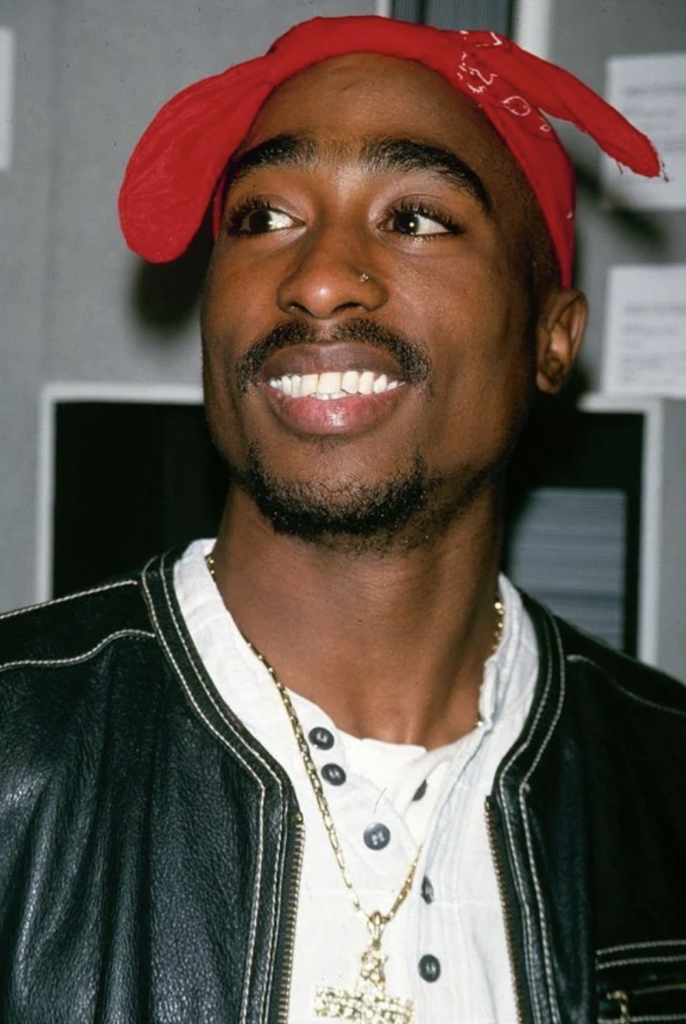 A man is smiling broadly while looking to the side. He has a red bandana tied on his head and is wearing a black leather jacket over a white shirt. He also has a gold chain necklace with a cross pendant. The background is out of focus and appears to be indoors.