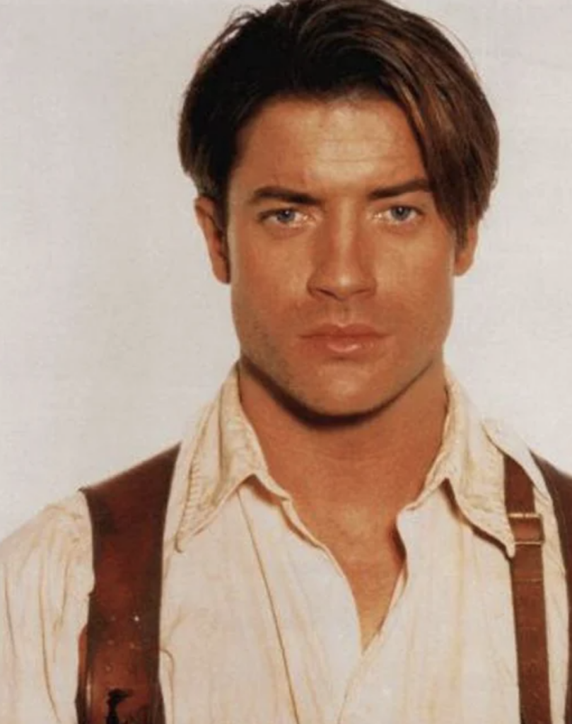 A person with short brown hair and blue eyes is facing the camera with a neutral expression. They are wearing a white shirt with a brown leather harness over it. The background is plain and light-colored.