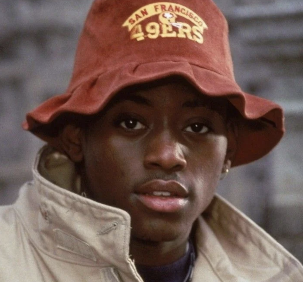 A young person wearing a red San Francisco 49ers bucket hat and a beige jacket looks towards the camera. The background is blurred, emphasizing the subject's face and attire.