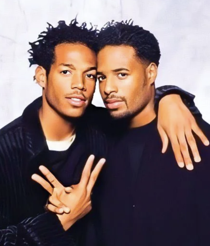 Two individuals pose together against a light background. The person on the left is making a peace sign with their right hand while the person on the right has their left arm around the other's shoulders. Both are wearing dark clothing and have short curly hair.