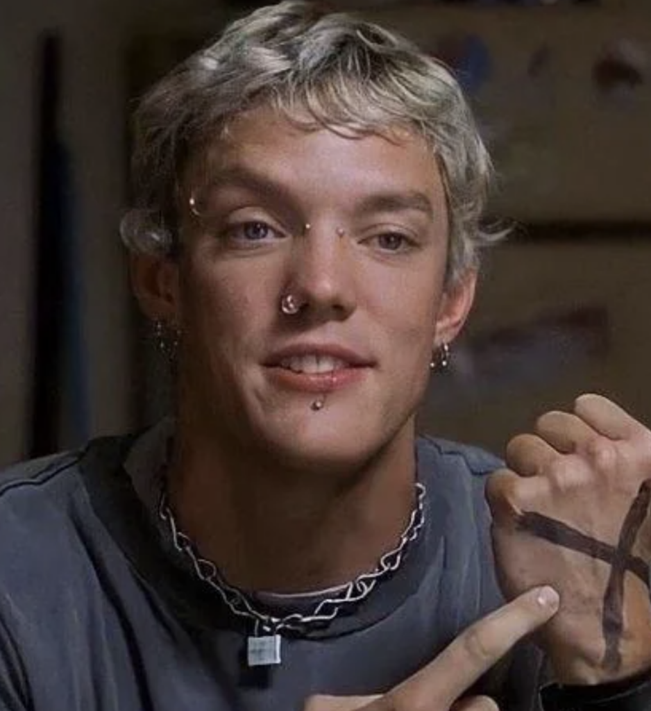 A person with short, light-colored hair and multiple facial piercings points to an X marked on their hand. They are wearing a dark T-shirt and a chunky chain necklace and have a neutral or slight smile on their face. The background is slightly blurred.
