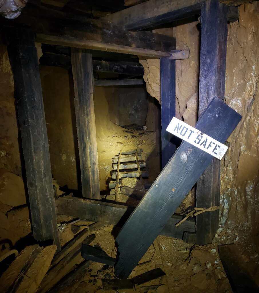 Dimly lit mine shaft with wooden beams and debris. A damaged sign labeled "NOT SAFE" is prominently displayed in the foreground, surrounded by fallen and broken wood. An unstable, narrow path leading further into darkness is visible in the background.