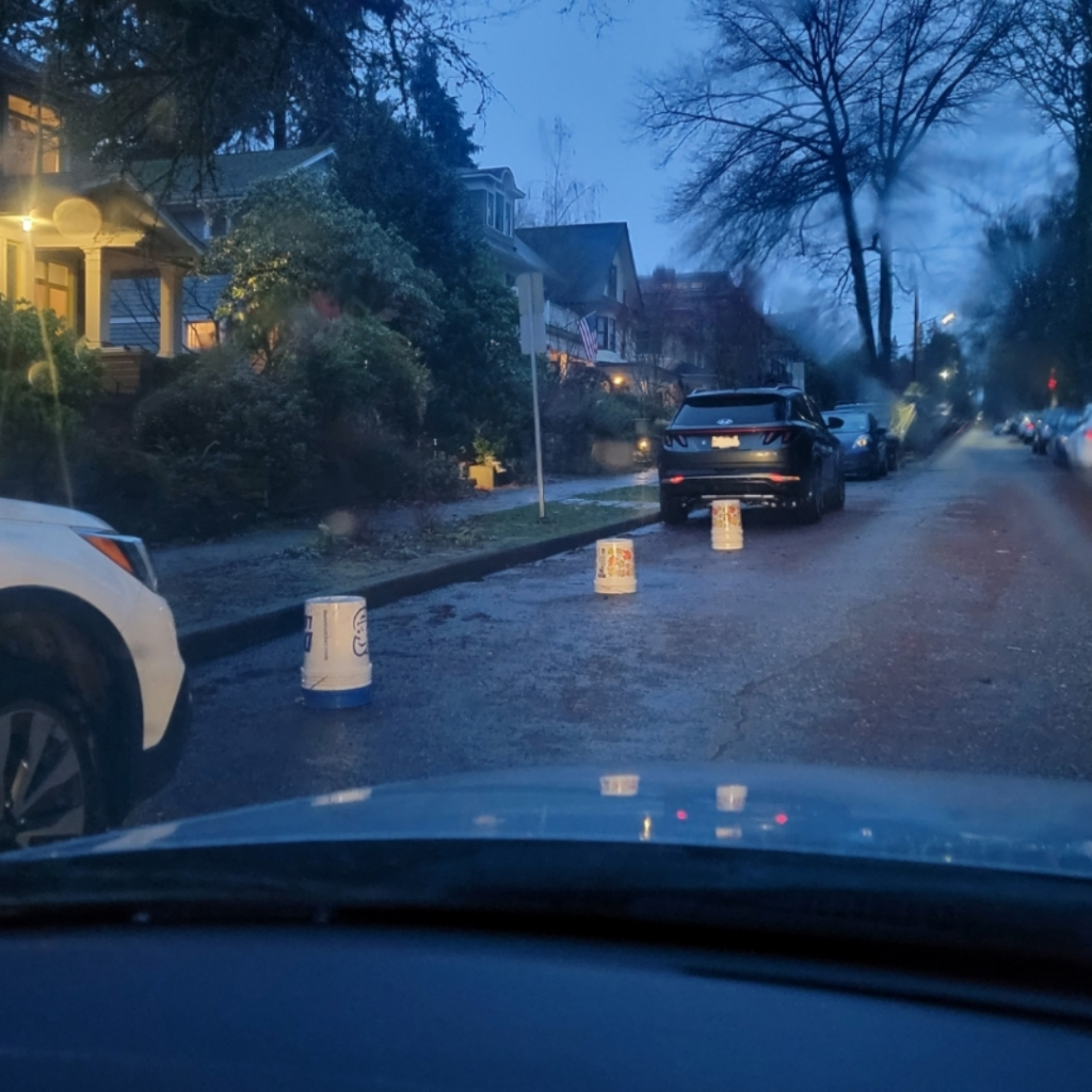 A residential street at dusk with several buckets placed on the road to reserve a parking space. There are houses and parked cars on either side, with trees lining the street. The scene is dimly lit, suggesting it might be evening.