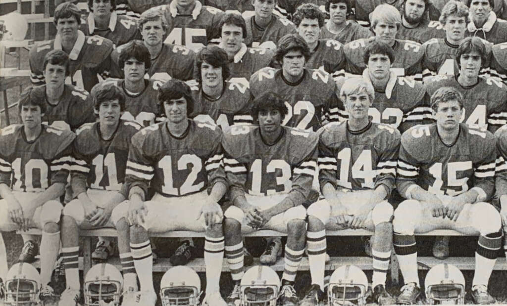 A black and white photo showing a group of young football players in uniform, sitting and standing in rows for a team picture. They are all wearing jerseys with numbers and football helmets are placed in front of them.