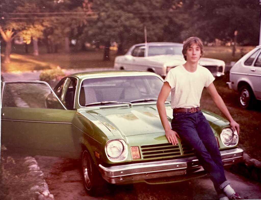 A young person with long hair sits on the hood of a green vintage car, wearing a white T-shirt and blue jeans. The driver's side door is open. There are two other cars, a white car and a light-colored car, parked nearby. Trees and grass are in the background.