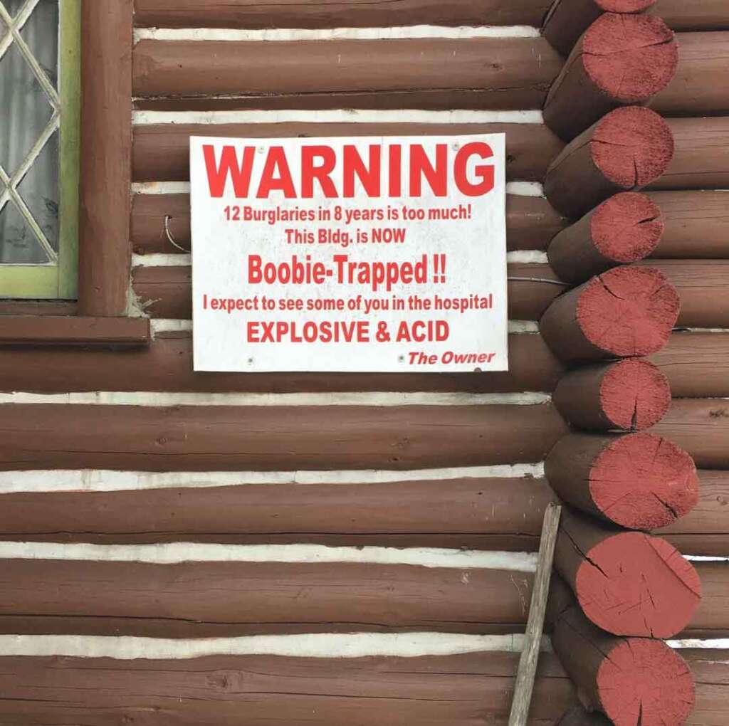 A warning sign on a log cabin wall reads: "WARNING. 12 Burglaries in 8 years is too much! This Bldg. is NOW Boobie-Trapped!! I expect to see some of you in the hospital. EXPLOSIVE & ACID. - The Owner.