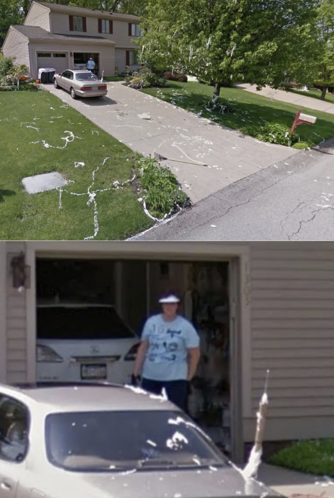 A photo of a residential driveway covered in toilet paper, spanning the yard, bushes, and a car. On the driveway, a person in shorts and a T-shirt is standing near the open garage, surveying the scene. Trees and a mailbox can be seen in the background.