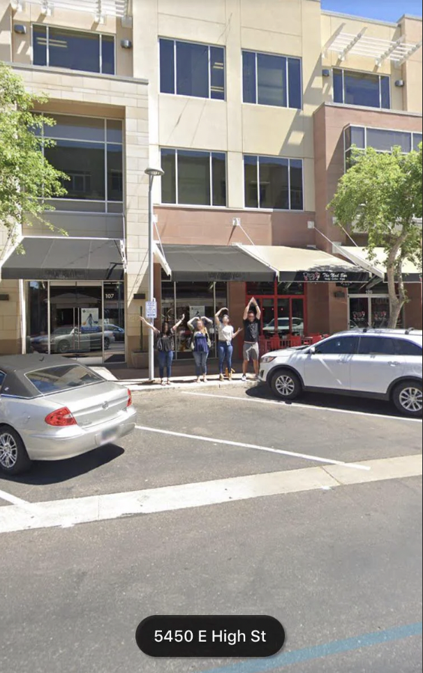 A group of people stand on the sidewalk in front of a modern, multi-story building with large windows and a red storefront. They are lined up near a parked white car and silver car. The scene is sunny, and trees are spaced along the sidewalk. The address "5450 E High St" is displayed.