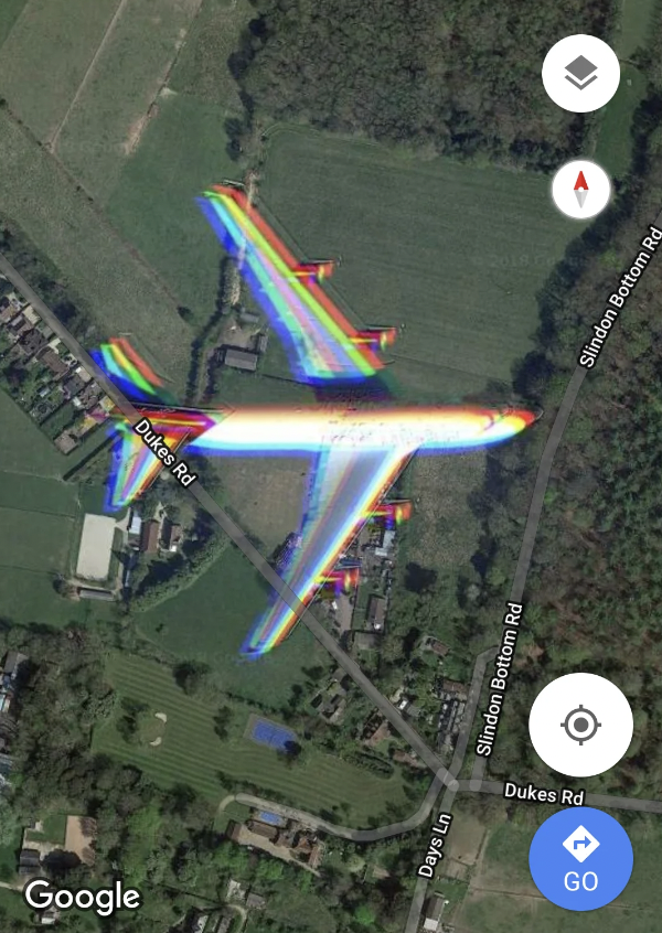 A satellite image shows a large airplane flying over a semi-rural area with fields, houses, and roads. The airplane appears to have a rainbow-colored blur effect, possibly due to image distortion. Nearby streets include Dukes Rd, Swindon Bottom Rd, and Days Ln.
