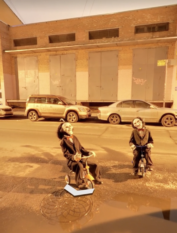 Two people wearing suits and eerie masks sit on tricycles in a dimly lit urban street. Behind them, a brick building with large garage doors and parked cars lines the street. The scene is spooky and surreal.