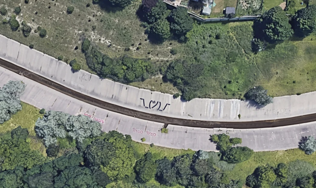 Aerial view of a curved concrete road bordered by lush green vegetation. The road has a visible dark line down its center and graffiti in large letters painted at various points along its surface. Trees and grassy areas are on either side of the road.