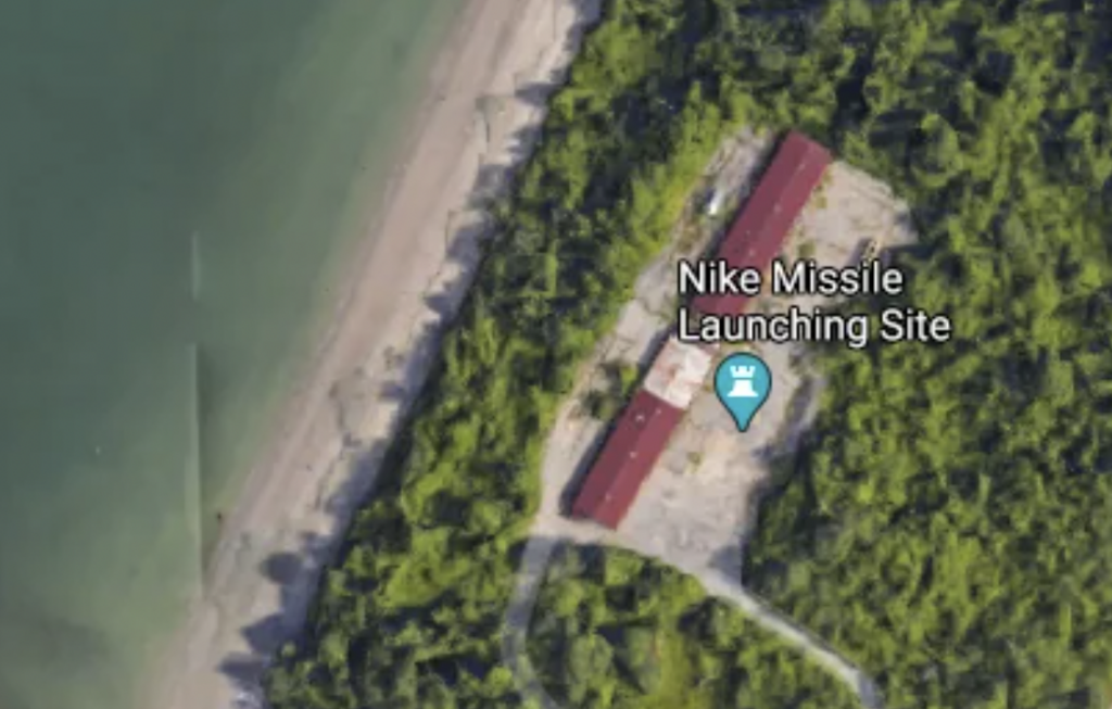 Aerial view of a forested area adjacent to a water body. In the middle of the greenery, there is a rectangular building labeled "Nike Missile Launching Site." The site is situated close to a shoreline, with a sandy beach visible along the water edge.
