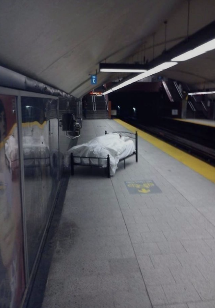 A neatly made bed, covered with a white blanket and pillows, is placed against a wall on a subway platform. The platform is lit with overhead lights, and there are tracks on the right. The bed appears unusual in this public transportation setting.