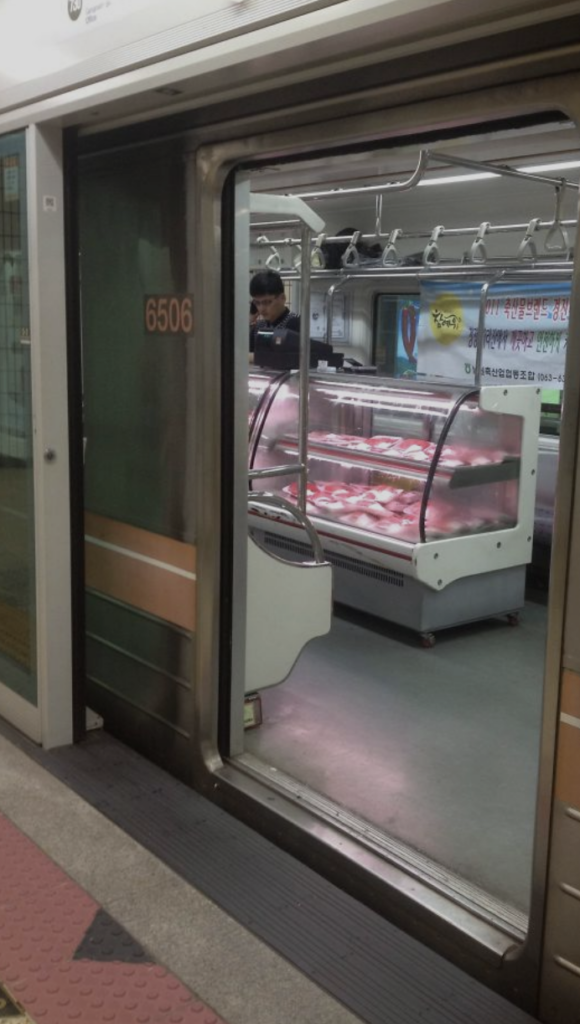 A subway door is open, revealing a meat vendor's stall inside the train car. The vendor stands behind a glass display case filled with various cuts of meat. The train car has overhead handrails and visible advertisements on the walls.