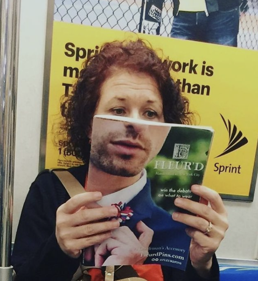 A curly-haired person with a dark sweater is sitting and reading a magazine. The cover of the magazine features a man's face, creating an amusing illusion where the reader’s eyes appear to align with the man's face on the cover. The background shows a Sprint advertisement.
