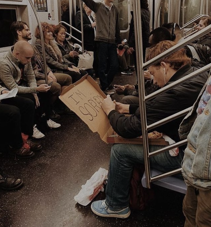 A person on a subway holds a large cardboard sign reading "I'M SORRY" as other passengers sit or stand around them. The scene shows people occupied with their activities, such as using their phones or conversing, in the subway car.