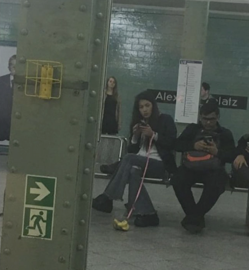 A woman and a man sit on a bench in a subway station, both looking at their phones. The woman has a pink leash attached to a banana on the ground. A green emergency exit sign and a structural column are in the foreground. A third person stands in the background.