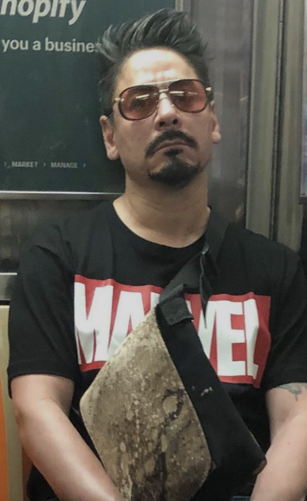 A man with spiked gray hair and a goatee is sitting on a subway. He wears sunglasses, a black T-shirt with a large red and white Marvel logo, and has a black crossbody bag with a beige front panel. His arms are crossed, and he appears to be looking ahead.