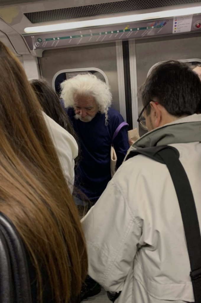 An older man with white, wild hair stands in a crowded subway car, looking down at something he is holding. He is surrounded by other passengers, some wearing coats and carrying bags. The interior of the subway car is visible, including the ceiling vents and door.