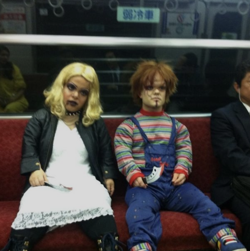 Two individuals, in costumes, sit on a subway. One is dressed as a blonde with a white dress and black jacket, resembling a horror character. The other is dressed as a doll in blue overalls and a striped shirt. Passengers sit nearby, some paying little attention.