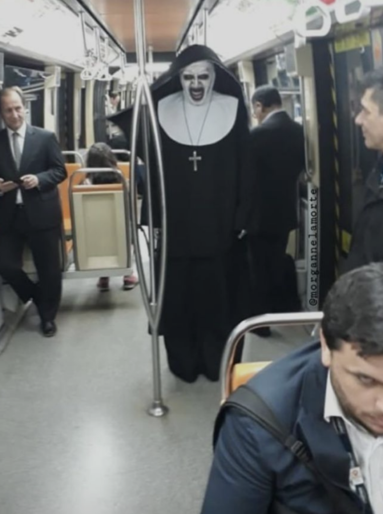 A person dressed in a scary nun costume with face paint and a cross necklace stands in the aisle of a subway train. The costume features a black habit and white wimple, resembling a character from a horror movie. Passengers around appear indifferent.